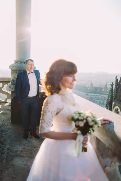 Bela noiva vestindo vestido branco posando na varanda velha, noivo no fundo — Fotografia de Stock