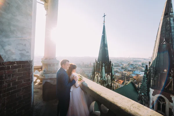 Schöner Bräutigam umarmt sinnliche Braut von hinten auf altem Balkon mit großartigem Stadtbild-Hintergrund — Stockfoto