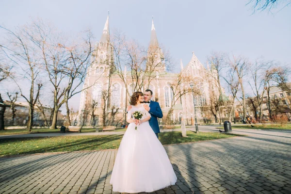 Handsome groom hugging sensual bride from behind  on background of an amazing gothic church — Stock Photo, Image