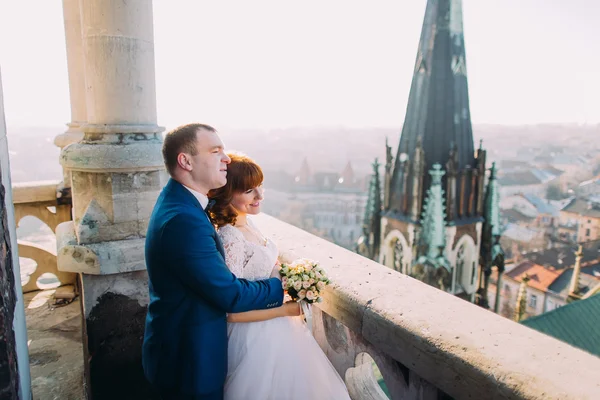 Bonito noivo sensual abraçando noiva recém-casada por trás na varanda do castelo velho com fundo da cidade — Fotografia de Stock