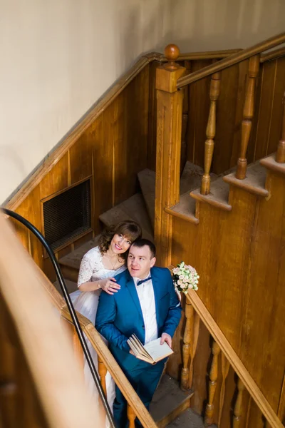 Vackra sensuella bruden kramas bakom sin stilig brudgum innehav bok på trätrappor av gamla bibliotek — Stockfoto