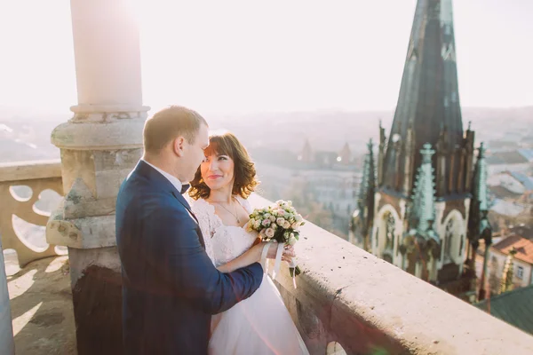 Schöner sinnlicher Bräutigam umarmt frisch vermählte Braut von hinten auf altem Burgbalkon mit Stadthintergrund — Stockfoto