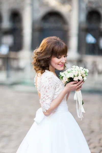Unga leende bruden tittar på ros bukett med stadsbilden i bakgrunden — Stockfoto