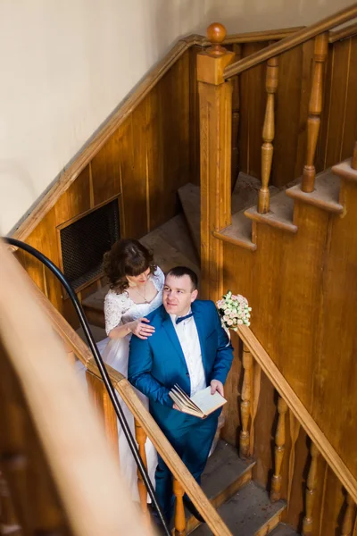 Прекрасная чувственная невеста обнимает из-за своего красивого жениха, держа книгу на деревянной лестнице старой библиотеки. — стоковое фото