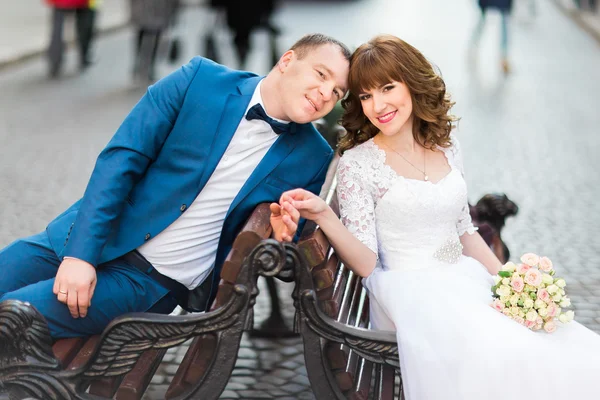 Casamento elegante noiva casal em vestido branco e elegante noivo sentado em um banco de mãos dadas — Fotografia de Stock