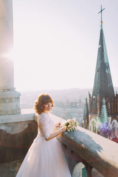 Schöne rothaarige Braut posiert mit Strauß auf altem Burgbalkon, Stadtbild Hintergrund — Stockfoto