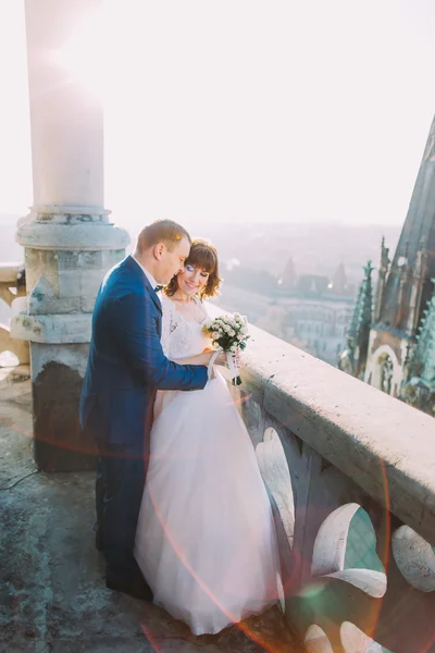 Beautiful couple, bride and groom posing on old balcony with column, cityscape background — Stock Photo, Image