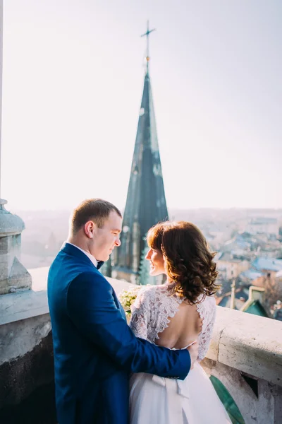 Gentle beautiful bride and groom holding hands with bouquet looking at each other on the ancient balcony — Stock Photo, Image