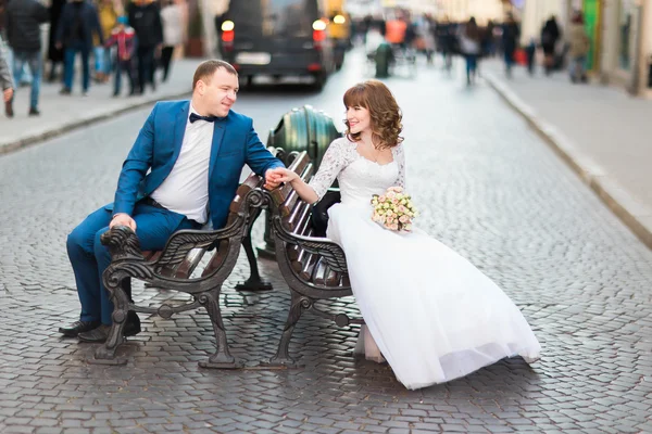 Casamento elegante noiva casal em vestido branco e elegante noivo sentado em um banco de mãos dadas — Fotografia de Stock