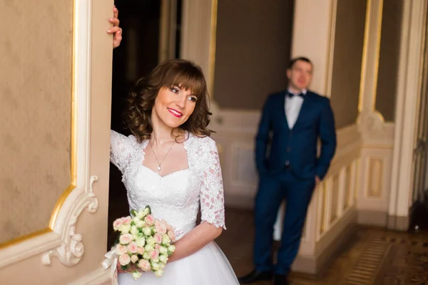 Noivo azulado fica atrás da noiva em vestido de renda com buquê de rosas em primeiro plano, dentro de casa — Fotografia de Stock