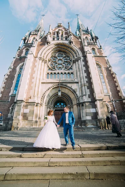 Noiva linda feliz e elegante noivo bonito de pé de mãos dadas no fundo de uma igreja de construção incrível — Fotografia de Stock