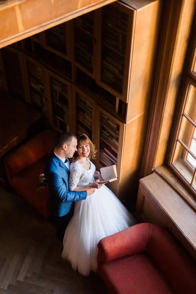 Casal de casamento elegante na biblioteca antiga. Noiva e noivo de pé livro de bruxas perto da janela. A vista de cima — Fotografia de Stock