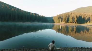 Handsome young thoughtful man in embroidery shirt throwing stones into the water and looking on the picturesque mountain lake Synevir. Ukrainian mythology concept. Back view