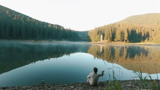 Handsome young thoughtful man in embroidery shirt throwing stones into the water and looking on the picturesque mountain lake Synevir. Ukrainian mythology concept — ストック動画