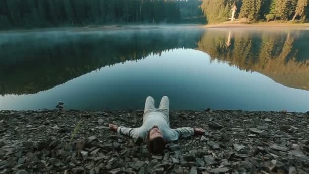 Pohledný mladý muž z tradičních výšivek ležících na skalnatém břehu vedle malebného horského jezera Synevir v Karpat. Okamžik harmonie mezi člověkem a přírodou — Stock video
