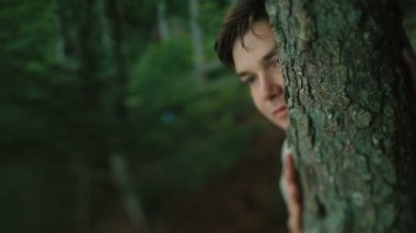Close up of young carpathian peasant watching someone in the mountain forest and hiding face by the tree