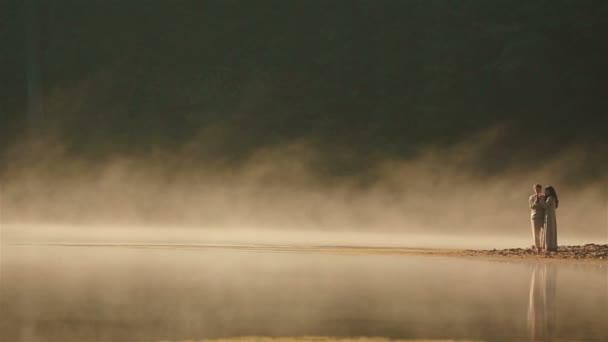 Couple de campagne en vêtements ethniques ukrainiens embrassant tendrement sur le rivage rocheux au coucher du soleil dans la brume dorée au pittoresque lac de montagne dans les Carpates. L'harmonie éternelle entre l'homme et la nature — Video