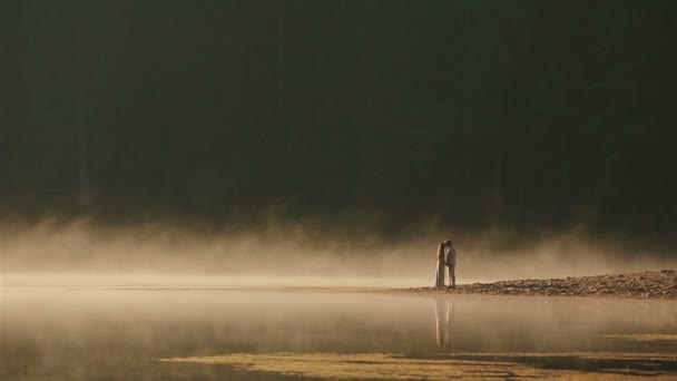 ウクライナの民族服の幸せな田舎のカップルは、カルパチアの美しい山の湖で黄金の霧の夕日に岩の海岸にキス。人間と自然の永遠の調和 — ストック動画