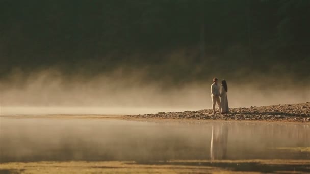 Сельская пара в украинской этнической одежде нежно обнимается на скалистом берегу на закате в золотом тумане у живописного горного озера в Карпатах. Вечная гармония между человеком и природой — стоковое видео
