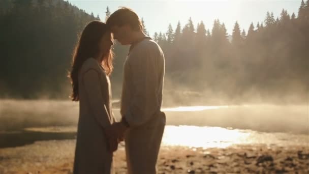 Heureux jeune couple de campagne en vêtements ethniques ukrainiens embrassant tendrement éclairé par les rayons du soleil sur le coucher du soleil dans la brume dorée au pittoresque lac de montagne dans les Carpates . — Video