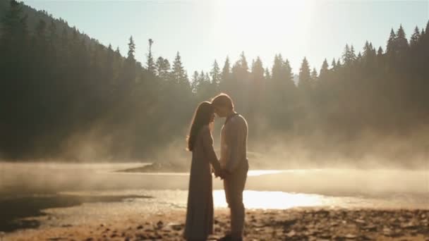 Breathtaking moment. Happy young countryside couple  in ukrainian ethnic clothes tenderly embracing lit by sunrays on sunset in golden mist at the picturesque mountain lake in Carpathians. — Wideo stockowe