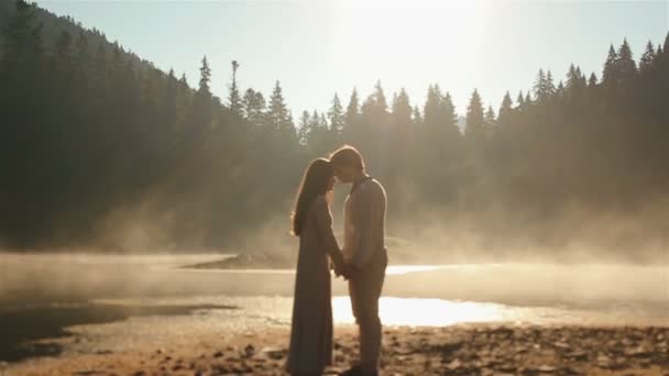 Momento de cortar a respiração. feliz jovem casal rural em roupas étnicas ucranianas ternamente abraçando iluminado por raios de sol no pôr do sol em névoa dourada no pitoresco lago da montanha em Cárpatos . — Vídeo de Stock