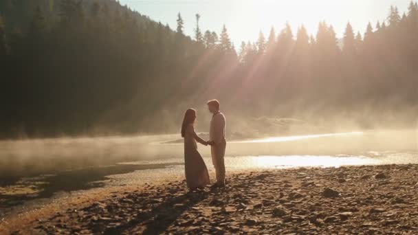 Momento impresionante. Feliz joven pareja de campo con ropa étnica ucraniana abrazándose tiernamente iluminada por los rayos del sol al atardecer en la niebla dorada en el pintoresco lago de montaña en los Cárpatos — Vídeos de Stock