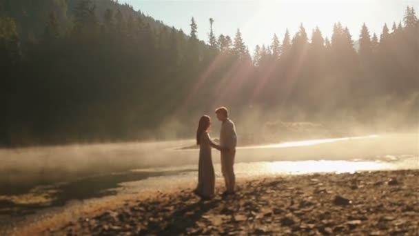 Momento de cortar a respiração. feliz jovem casal rural em roupas étnicas ucranianas ternamente abraçando iluminado por raios de sol no pôr do sol em névoa dourada no pitoresco lago da montanha em Cárpatos . — Vídeo de Stock