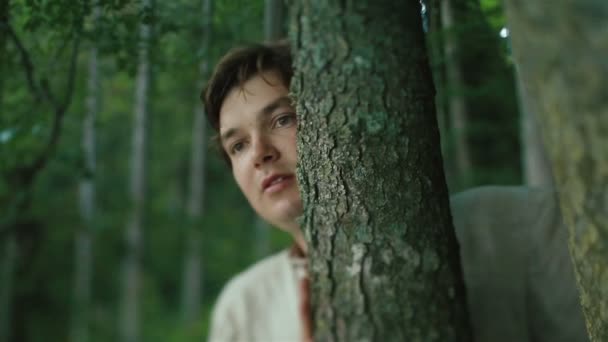 Крупним планом молодий карпатський селянин спостерігає за кимось у гірському лісі і ховається біля дерев — стокове відео