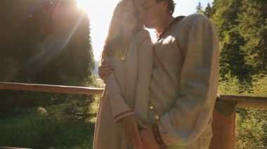 Beautiful young couple in traditional ukrainian clothes softly kissing on the wooden bridge in mountains. Majestic Carpathians on background. Very tender moment