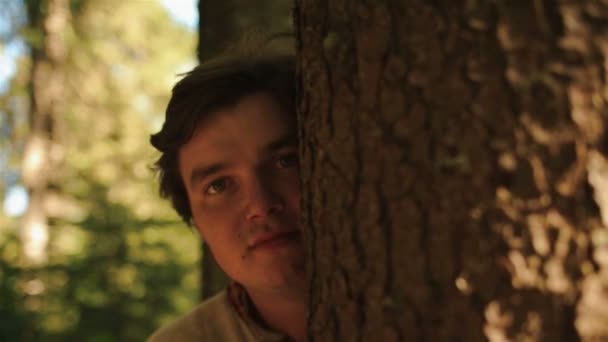 Крупным планом загипнотизированного молодого человека, наблюдающего за кем-то в лесу и закрывающего лицо деревом. История любви в горах — стоковое видео