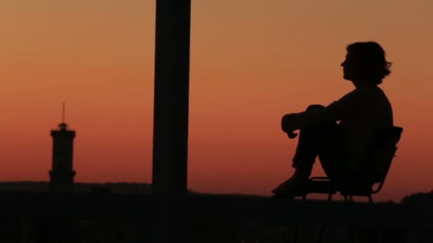 Sylwetka smutny młoda kobieta siedzi na balkonie z tło miasta na zachód słońca — Wideo stockowe