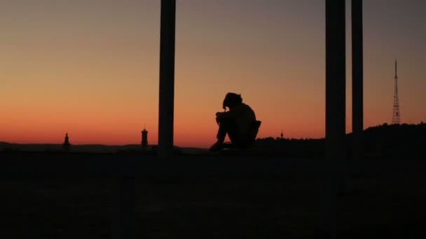 Silhouette of sad thoughtful young woman sitting on the balcony with background of the city on sunset — Stock Video