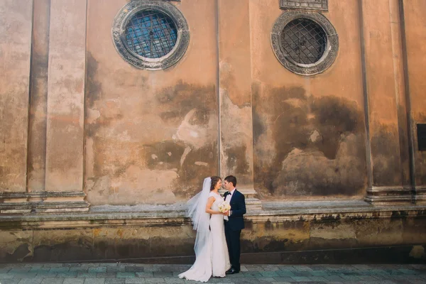 Linda noiva em vestido branco e belo noivo segurando buquê de noiva cara a cara perto da parede marrom da igreja velha — Fotografia de Stock