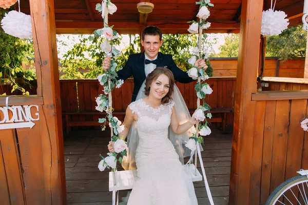 Retrato de belo noivo em pé atrás da bela noiva sentada no balanço — Fotografia de Stock