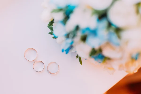 Close up de três anéis de casamento dourados com belo buquê floral no fundo branco — Fotografia de Stock