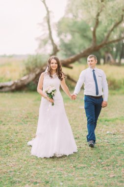 Yakışıklı damat ve gelin beyaz peçe geri arka plan yeşil orman üzerinde el ele tutuşarak yürüyüş