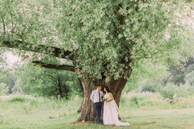 Mutlu genç gelin ve damat taze yeşil yaprakları ile büyük bir ağaç altında poz çayır bahar