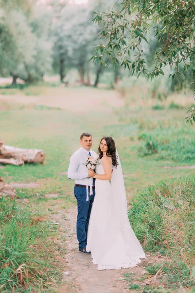 Apuesto novio y novia en velo blanco de pie tomados de la mano en el bosque verde de fondo — Foto de Stock