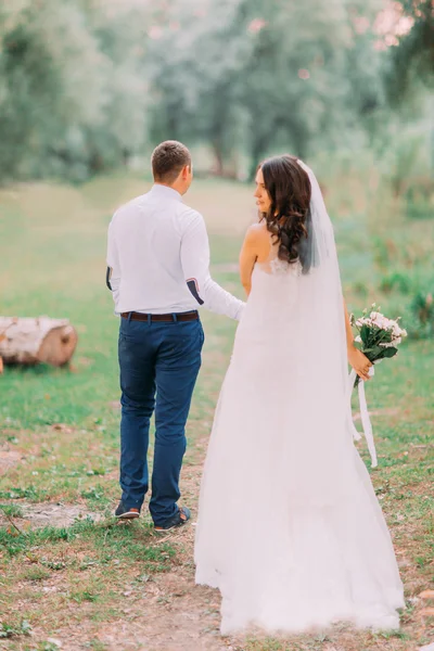 Guapo novio y novia en velo blanco caminando de espaldas tomados de la mano en el bosque verde de fondo — Foto de Stock