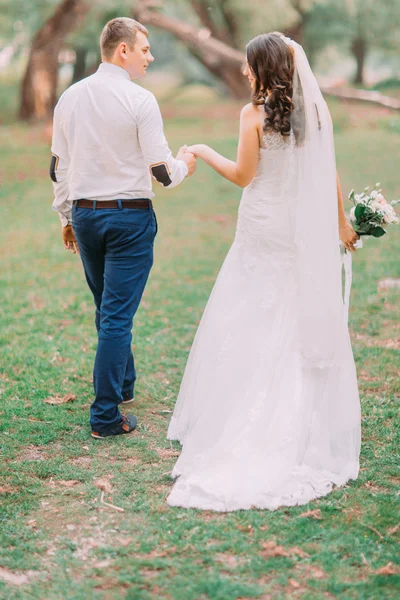 Guapo novio y novia en velo blanco caminando de espaldas tomados de la mano en el bosque verde de fondo — Foto de Stock