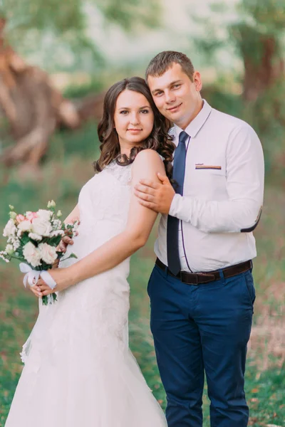 Красивая невеста с свадебным букетом и женихом позирует перед камерой на фоне зеленого парка — стоковое фото