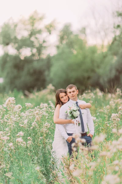 Beautiful bride embracing from behind her handsome groom in the summer meadow with wild flowers — Stock Photo, Image