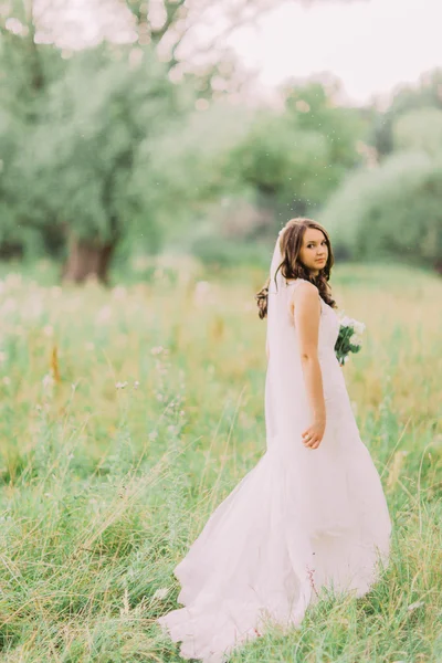 Potret pengantin muda yang cantik dengan gaun yang elegan dengan kerudung panjang dan buket bunga putih di taman — Stok Foto