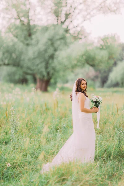 Porträt der schönen jungen Braut in elegantem Kleid mit langem Schleier und weißem Blumenstrauß im Park — Stockfoto