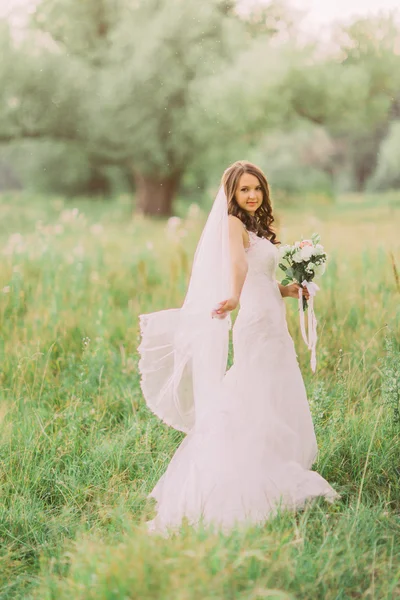 Porträtt av vackra unga brud i elegant klänning med lång slöja och vita blommor bukett i parken — Stockfoto