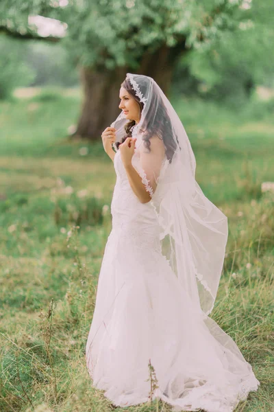 Cantik pengantin dalam gaun putih bermain dengan kerudung, taman musim semi hijau di latar belakang — Stok Foto