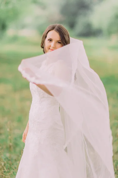 Cantik pengantin dalam gaun putih bermain dengan kerudung, taman musim semi hijau di latar belakang — Stok Foto