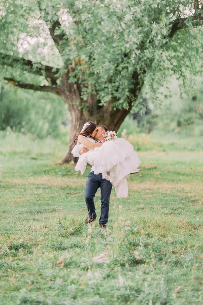 Молодий усміхнений наречений носить свою красиву дружину в весняному зеленому парку — стокове фото