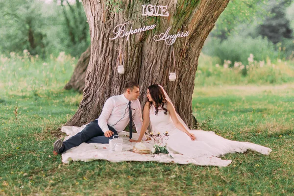 Schönes Hochzeitspaar sieht einander sitzend auf Plaid an, Picknick unter Baum mit stilvollem Dekor — Stockfoto